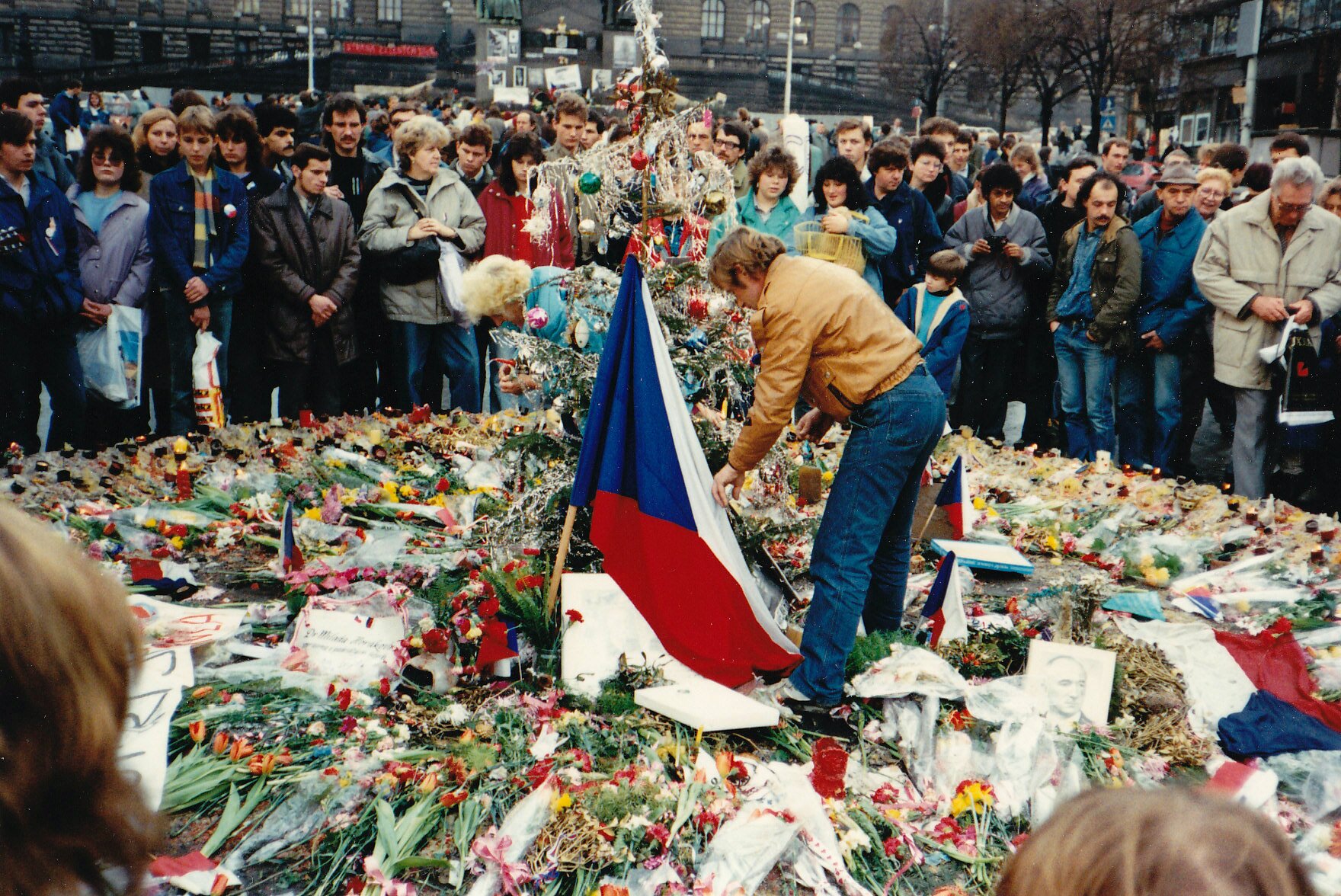 Zdjęcie. Na ziemi leżą stosy kwiatów. Po środku stoi mężczyzna i układa kwiaty obok czechosłowackiej flagi. Wokół stoją ludzie w jesiennych ubraniach. W tle kamienice i domy.
