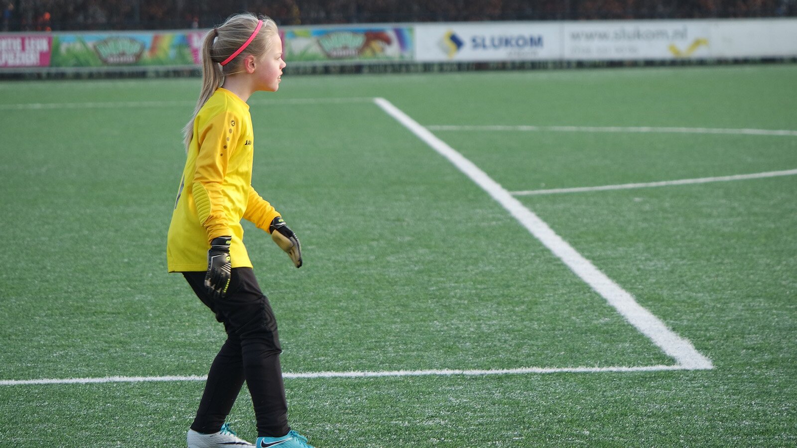 Zdjęcie przedstawia dziewczynkę stojącą bokiem na murawie boiska, zwróconą w prawo. Dziewczynka ubrana jest w strój bramkarza, czyli długie czarne spodnie, żółtą bluzkę z długim rękawem oraz rękawice bramkarskie.
