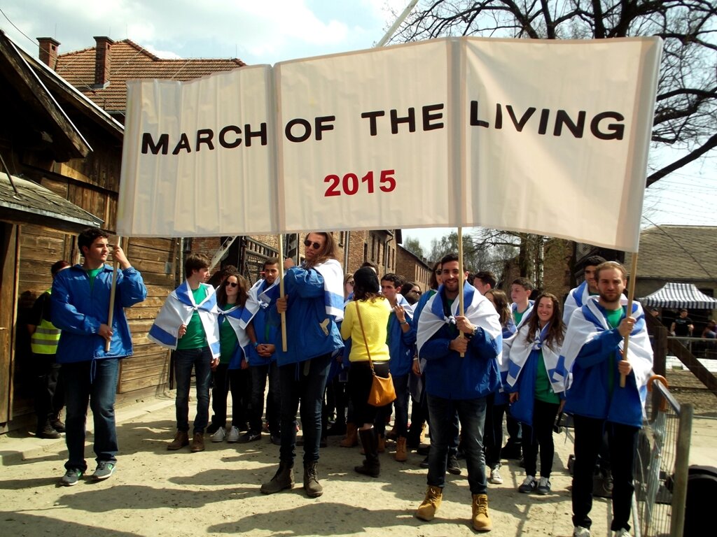 Fotografia przedstawiająca manifestujących ludzi, którzy niosą transparent z napisem "March of the living dwa tysiące piętnasty". Marsz odbywa się na terenie byłego niemieckiego obozu zagłady Auschwitz. 