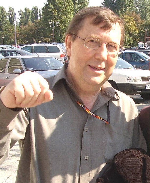 Kliknij, aby powiększyć Fotografia przedstawia mężczyznę w średnim wieku. Ma okulary, ciemne włosy z wysokimi zakolami. Ma na sobie szarą koszulę, na szyi wisi mu smycz reklamowa, której koniec znajduje się w kieszonce koszuli. Podnosi do góry zaciśniętą prawą pięść, w geście "żółwika". Widać go od pasa w górę. Za nim znajdują się zaparkowane samochody, a dalej drzewa.