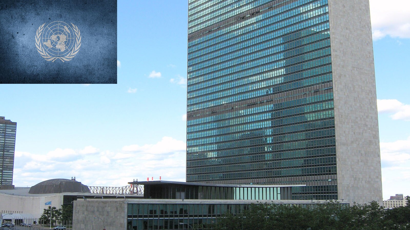Zdjęcie przedstawia po prawej stronie wysoki, kilkudziesięciopiętrowy budynek, którego dłuższy bok jest cały przeszklony. Poniżej widoczne są niższe budynki wykonane z betonu i szkła, w tym jeden z niewielką kopułą. W lewym górnym rogu widoczna jest flaga Organizacji Narodów Zjednoczonych. Na niebieskim tle widoczna jest mapa świata z poszczególnymi kontynentami, oglądana z czterdziestego równoleżnika szerokości południowej, na setnym stopniu szerokości zachodniej od Greenwich. Mapa otoczona jest dwiema gałązkami oliwnymi.