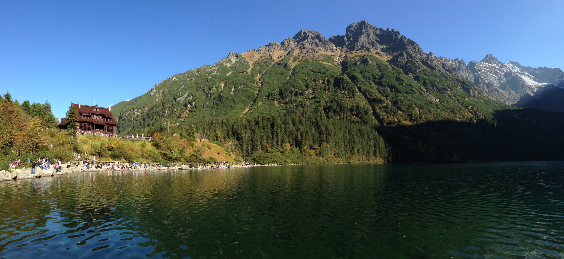 Zdjęcie przedstawia Morskie Oko w Tatrach. Zostało ono wykonane znad powierzchni jeziora. Woda ma niebieską barwę, na dalszym planie widać kamienisty brzeg jeziora. Stoi tam wielu turystów. Na drugim brzegu widoczna jest roślinność: krzewy oraz liczne drzewa iglaste. W lewym rogu zdjęcia znajduje się schronisko nad Morskim Okiem - budynek wykonany z ciemnego drewna. Za lasem iglastym widoczne są tatrzańskie, skaliste szczyty, w dolnych partiach porasta je roślinność. 