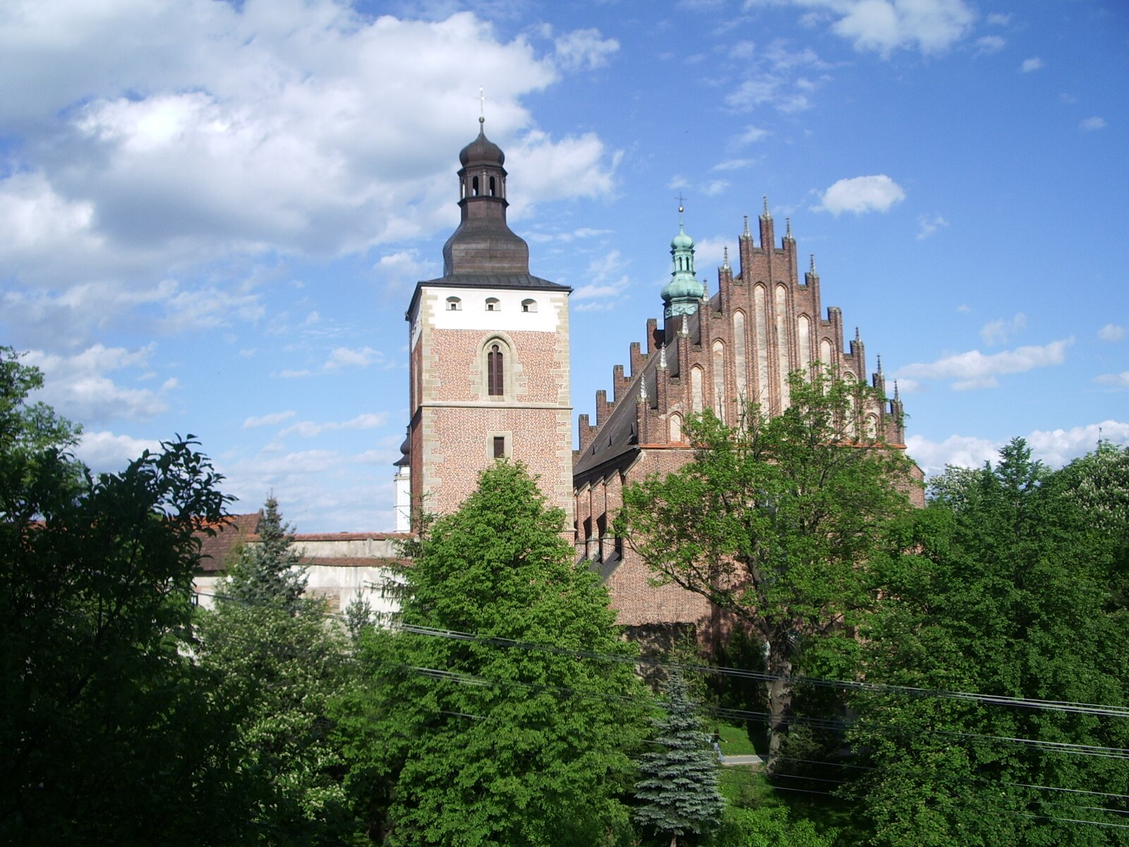 Fotografia przedstawia ceglany budynek ze strzelistym dachem i wieżą stojący pośród drzew. Na szczycie wieży widać krzyż.
