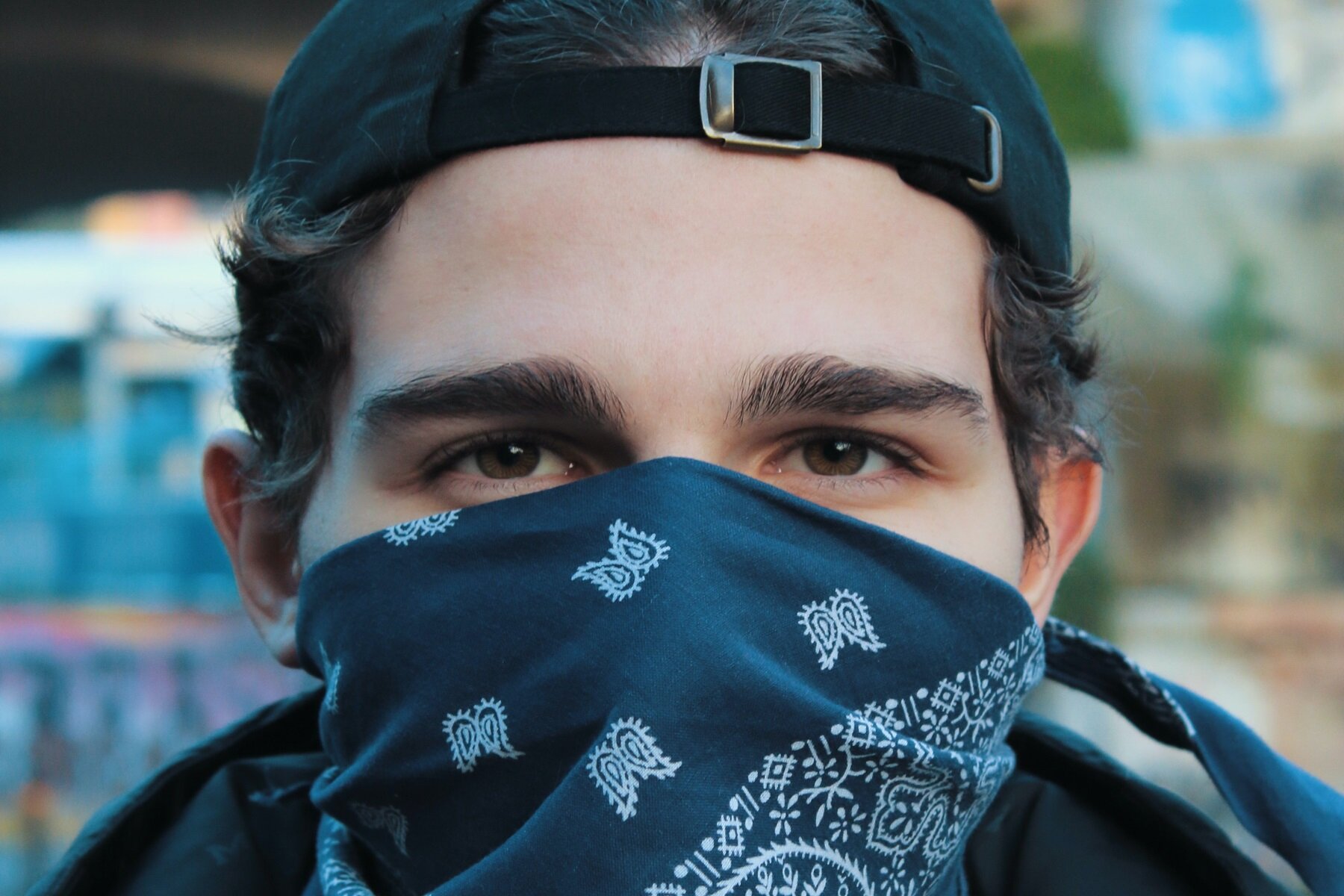 Zdjęcie przedstawia głowę młodego chłopaka z twarzą skierowaną w obiektyw. Chłopiec ma ciemne, błyszczące oczy. Dolna część twarzy jest zasłoniętą czarną chustą, tak zwaną bandaną, w białe wzory. Chłopiec ma ciemne, krótkie włosy, a na głowę założoną odwróconą czapkę z daszkiem. Tło zdjęcia jest rozmyte.