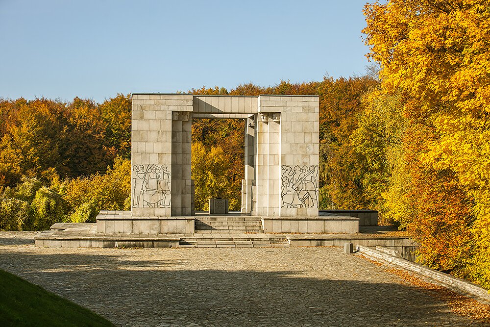 Zdjęcie przedstawia pomnik. Jest to łuk z piaskowych bloków, na którego elewacji znajdują się wyrzeźbione postaci. Po lewej są to mężczyźni z kijem i kosą, a po prawej żołnierze z bagnetem, którzy uderzają bezbronną kobietę. W tle rosną drzewa.