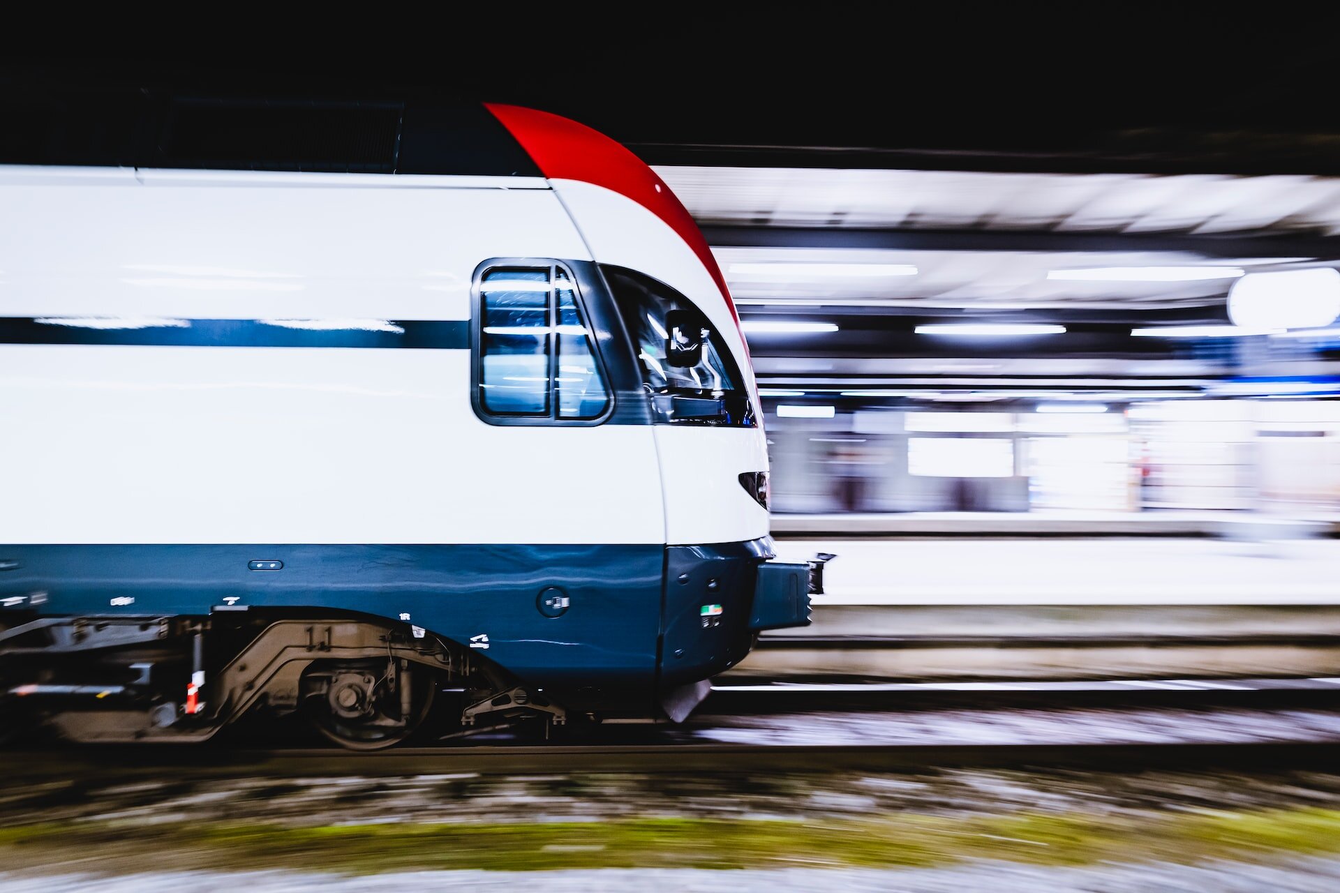 Zdjęcie przedstawia chwilę, gdy nowoczesny pociąg wjeżdża na peron dworca, a w tle widoczne są jasno oświetlone przestrzenie.