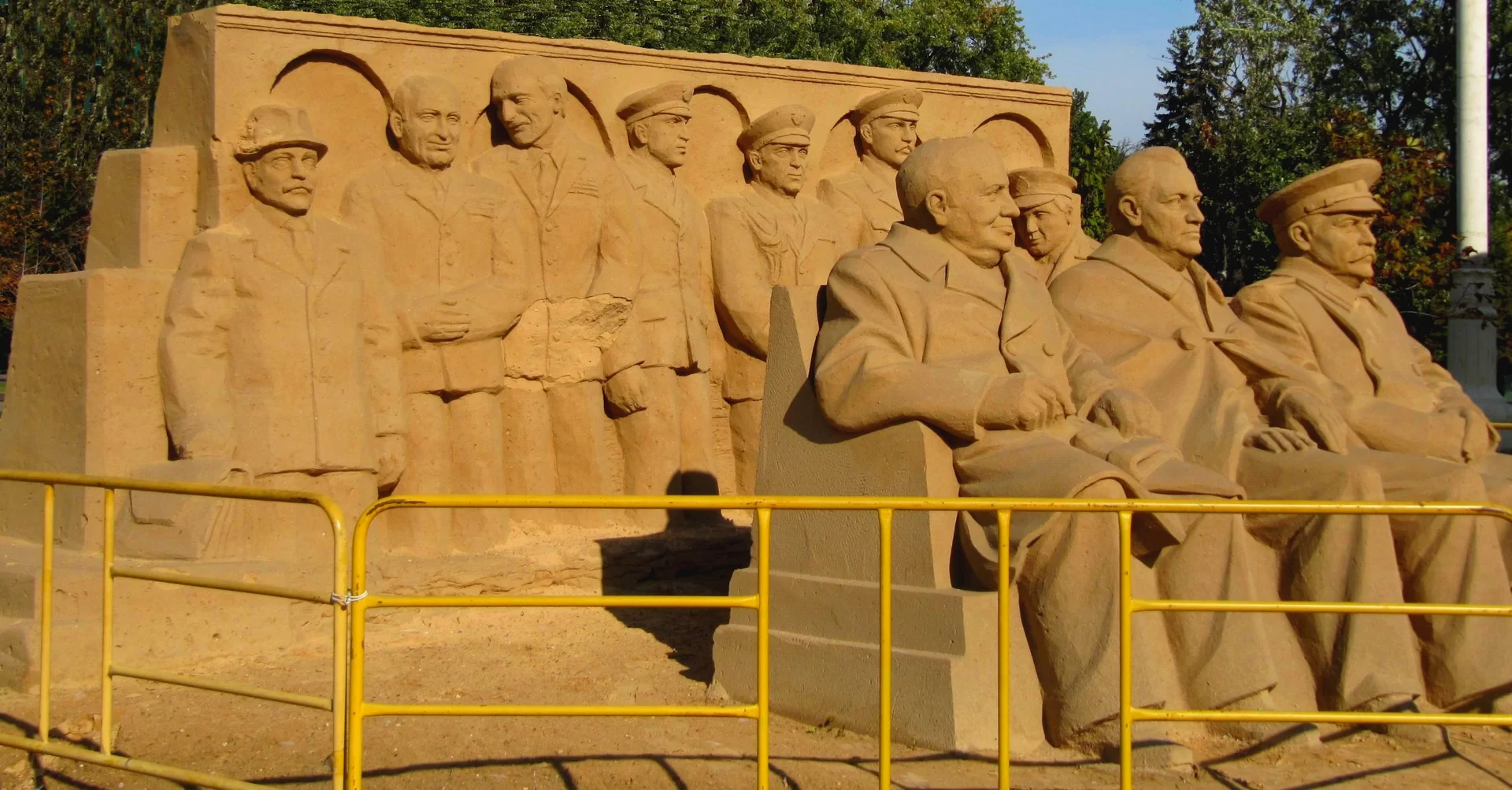 Kliknij, aby powiększyć Zdjęcie przedstawia kamienny pomnik, na którym obok siebie siedzą: Churchill, Roosevelt i Stalin. Za nimi stoi siedmiu mężczyzn.