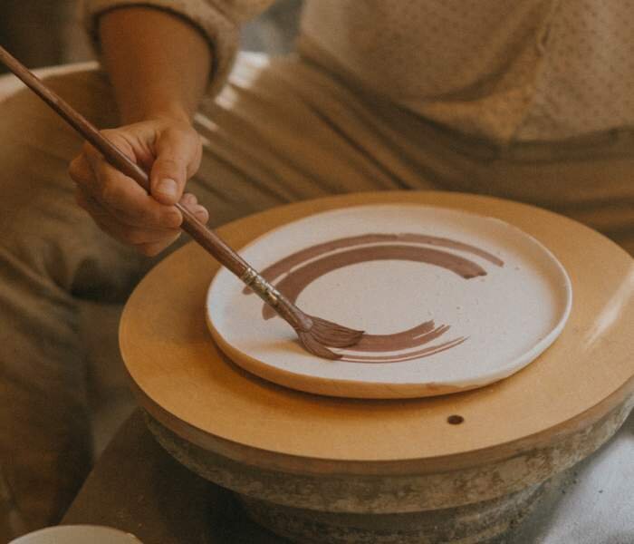 Wskazywanie surowców do przygotowania szkliw ceramicznych. Pierwsze zdjęcie przedstawia osobę malującą za pomocą pędzla gliniany talerz.