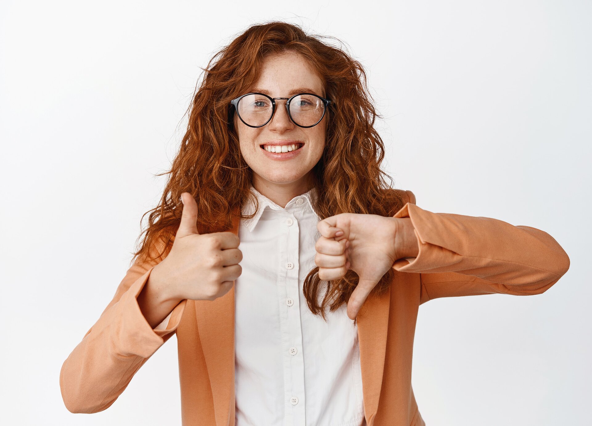 Zdjęcie przedstawia uśmiechniętą młodą kobietę, w ujęciu od pasa w górę. Kobieta ma długie, rude, lekko pofalowane włosy, ubrana jest w białą koszulę, jasnobrązową marynarkę oraz ma okulary korekcyjne. Obie ręce ma podniesione na wysokość klatki piersiowej. Lewa dłoń ma kciuk skierowany w dół, a prawa dłoń ma kciuk skierowany do góry. 