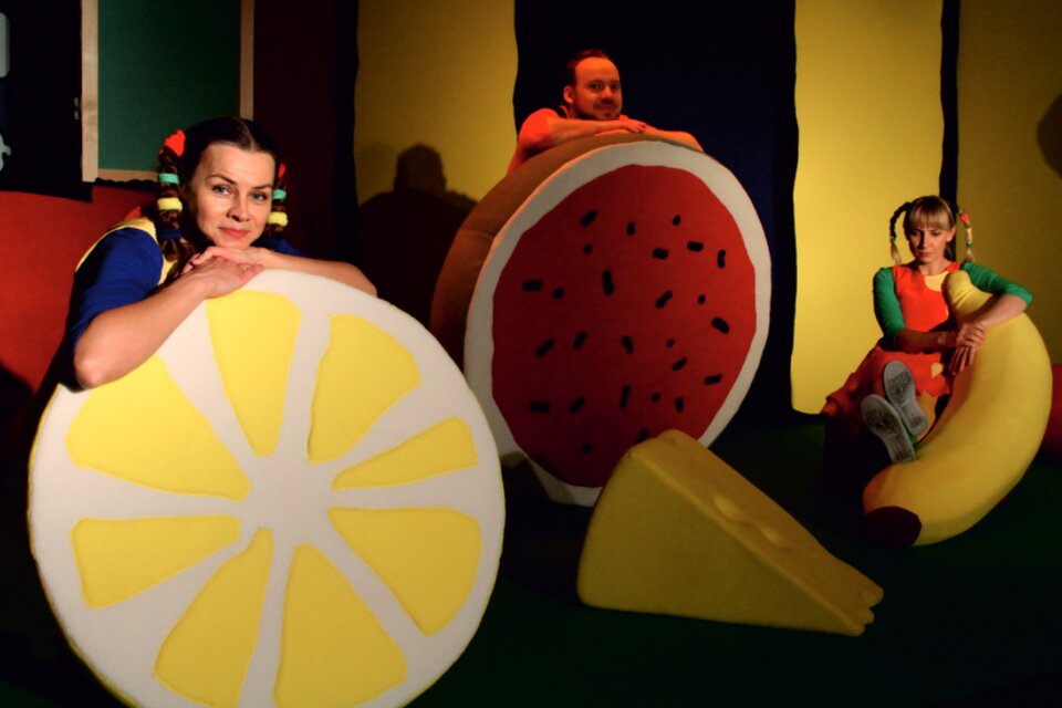 Ilustracja przedstawia trzy osoby opierające się o rekwizyty sceniczne. Po lewej dziewczyna z kitkami opiera się o połówkę cytryny. Mężczyzna dalej oparty jest o arbuz, a dziewczyna z prawej siedzi oparta o dużego banana.