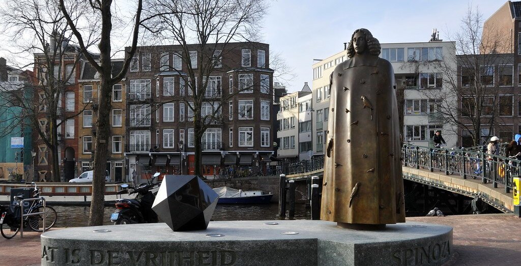 Zdjęcie przedstawia plac, na na nim stojący pomnik mężczyzny w sięgającej stóp pelerynie. Mężczyzna jest młody, ma włosy sięgające ramion. W tle rzeka i niewielki most. Na drugim brzegu liczne budynki mieszkalne.