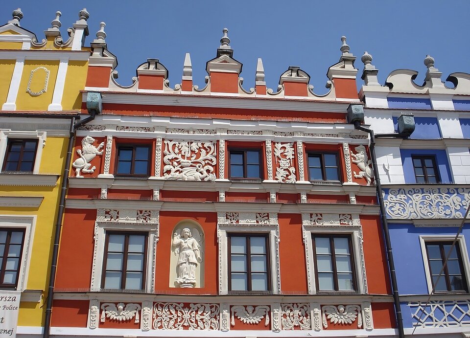 Zdjęcie przedstawia widok współczesny na górną partię jednej z kamienic ormiańskich na rynku w Zamościu. Kamienica o czerwonym kolorze ściany, prostokątnych oknach, jest dzielona kondygnacjami na fasadzie przez gzymsy i fryzy. Na szczycie fasady renesansowa attyka osłaniająca dach, zdobiona sterczynami i wolutami. Dominują zdobnicze motywy roślinne i zwierzęce na fryzach. Pośrodku jednej z kondygnacji, piętra uchwyconego bliżej dolnej krawędzi zdjęcia, widoczna we wnęce figura postaci. Po obu stronach czerwonej kamienicy fragmenty attyk i ścian kamienic sąsiednich - żółtej z lewej strony i błękitnej z prawej.