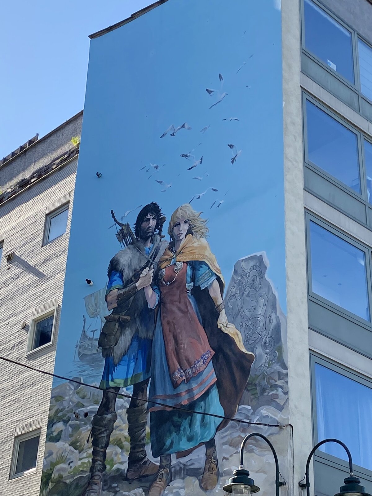 Na zdjęciu znajduje się namalowana na bocznej ścianie bloku kobieta ubrana w niebiesko‑czerwoną suknię oraz mężczyzna ubrany w niebieski strój oraz przepasaną zwierzęcą skórę. W tle znajdują się skały oraz jezioro na którym zacumowana jest łódka.