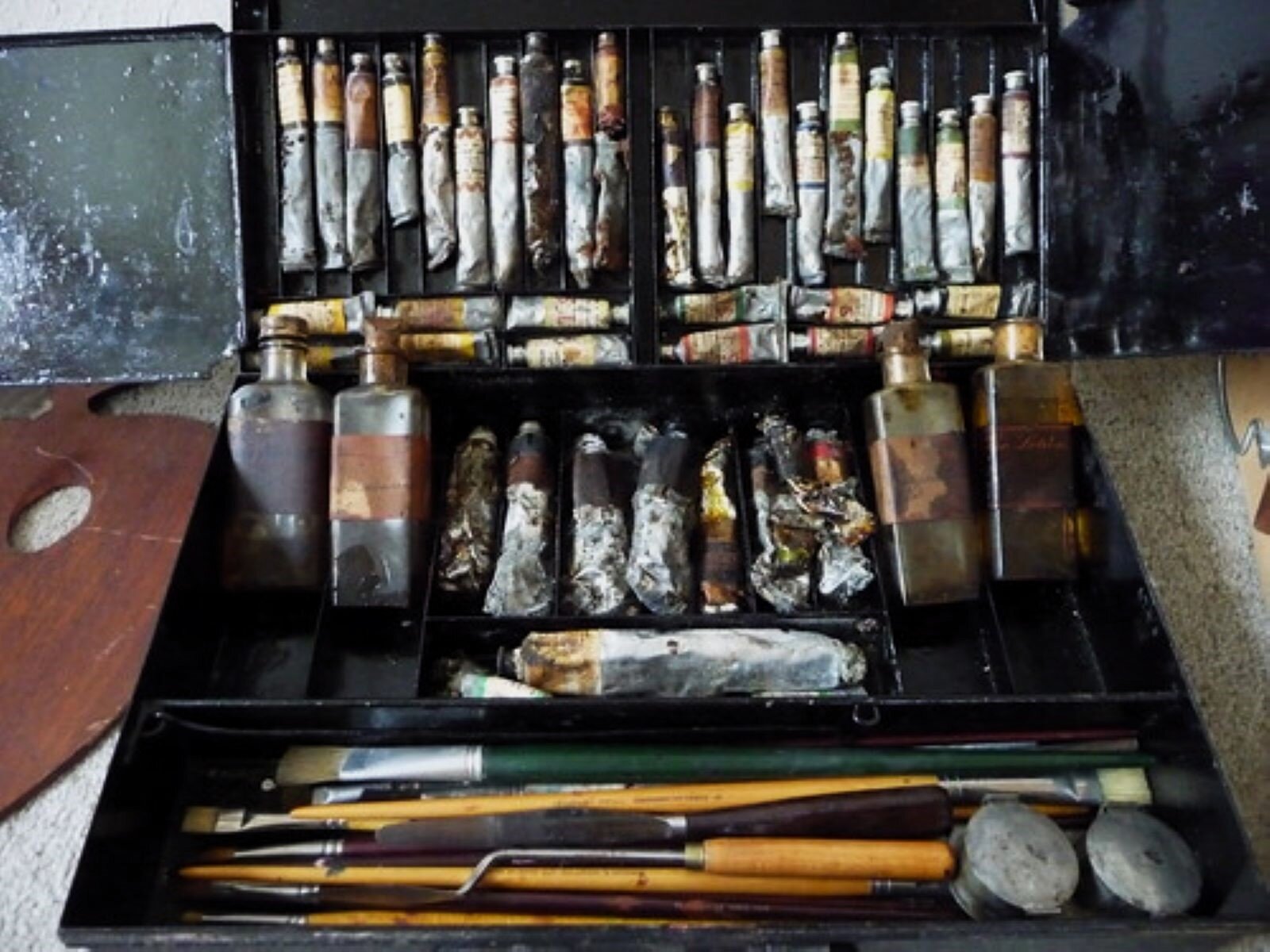 Zdjęcie przedstawia otwartą kasetę malarską z farbami, pędzlami i pojemnikami z olejem i terpentyną. Na wiek stoją poukładane zakrętkami do góry wąskie tubki z farbą. Są one pobrudzone i widać, że farba była z nich wielokrotnie wyciskana. Pod nimi leżą ułożone pionowo dwa rzędy farb. W drugiej części kasetki leży osiem ułożonych jedna obok drugiej większych tub z farbą. Z prawej i lewej strony tych tub stoją po dwie szklane butelki z terpentyną. Najbliżej widza, na pierwszym planie w poziomej przegródce leżą różnej wielkości i długości pędzle oraz szpachelka. W prawym rogu dwa okrągłe pojemniki z cyny do rozmieszana farb.