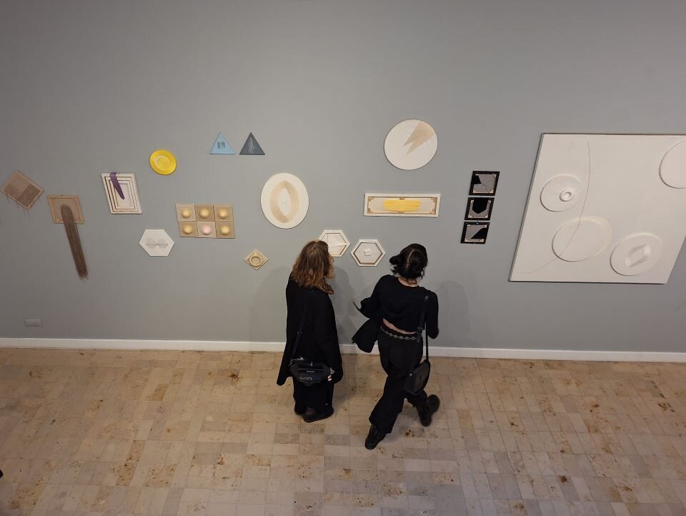 Ilustracja przedstawia fotografię wnętrza wystawy malarstwa w galerii. Na ścianie wiszą dzieła różnych rozmiarów i w różnych formatach. Są rozwieszone na różnych wysokościach i w rożnych odstępach. Na pierwszym planie znajdują się dwie postaci zwiedzające wystawę i przyglądające się wiszącym pracom. Sylwetki są ukazane od tyłu. Fotografia jest wykonana z perspektywy z lotu ptaka.