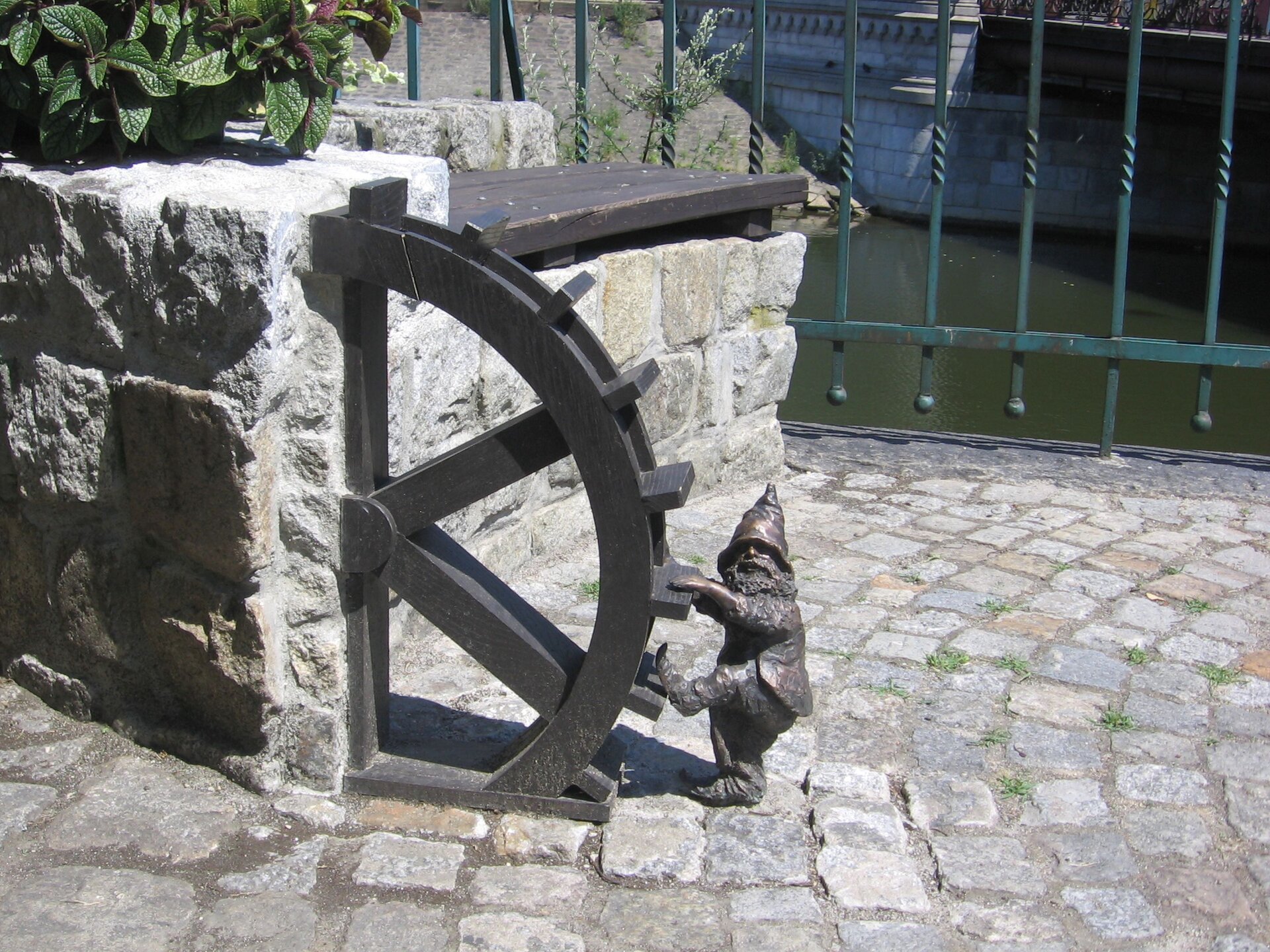 Ilustracja przedstawia rzeźbę jednego z wrocławskich krasnali o imieniu Młynarz. Jest to krasnal w czapce i z brodą, który próbuje wejść na koło młynarskie. Lewa ręka i lewa noga są uniesione w górę i wygląda tak, jakby krasnal wchodził po drabinie. Rzeźba stoi na brukowanej powierzchni. W tle widać barierki i rzekę.