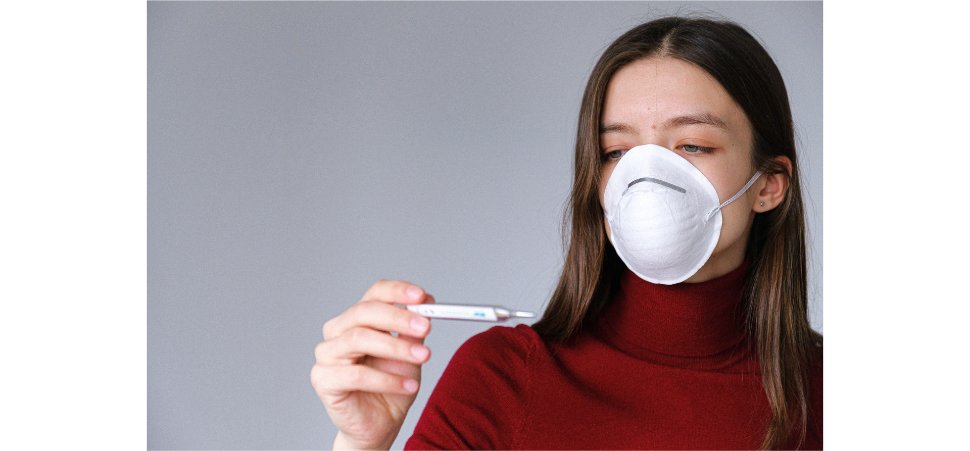 Zdjęcie przedstawia kobietę z długimi włosami w golfie. Kobieta ma na twarzy maseczkę ochronną. Kobieta spogląda na termometr, który trzyma w ręku.