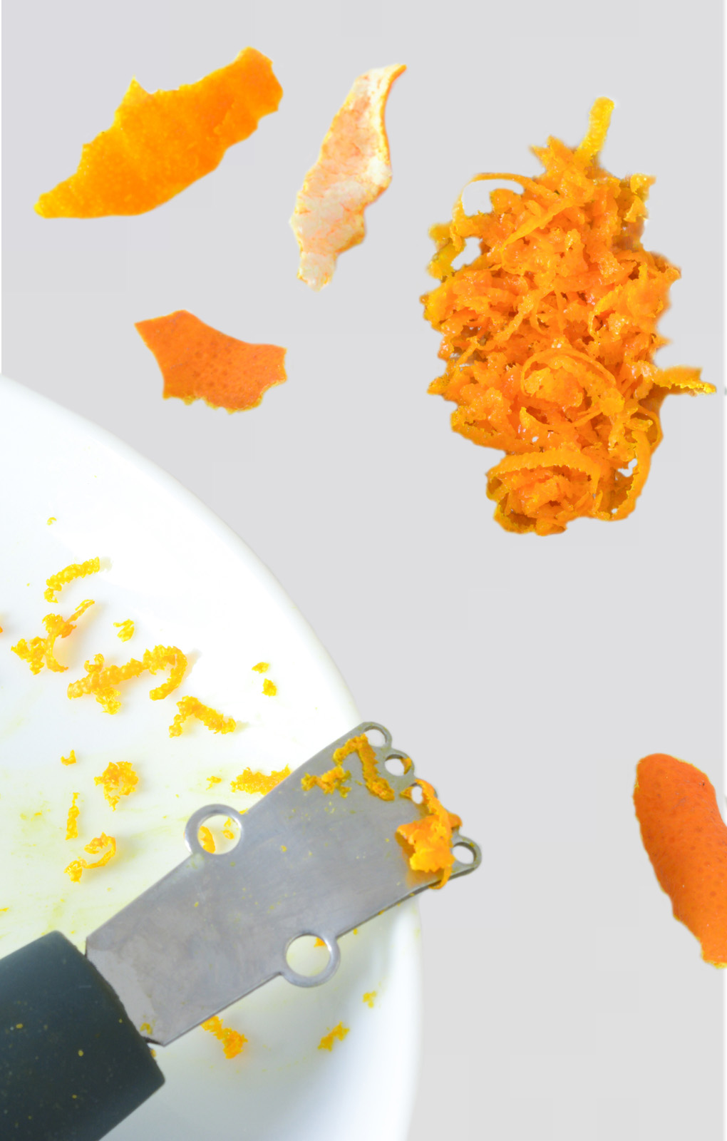 Na zdjęciu widać zester, przyrząd służący do uzyskiwania startej skórki w postaci cieniutkich pasków z cytryny lub innych owoców cytrusowych. Zester składa się z czarnego uchwytu i zakrzywionego metalowego końca, którego górna część ma ostre oczka. Obok zestera leży starta skórka z pomarańczy w postaci cieniutkich pasków. 