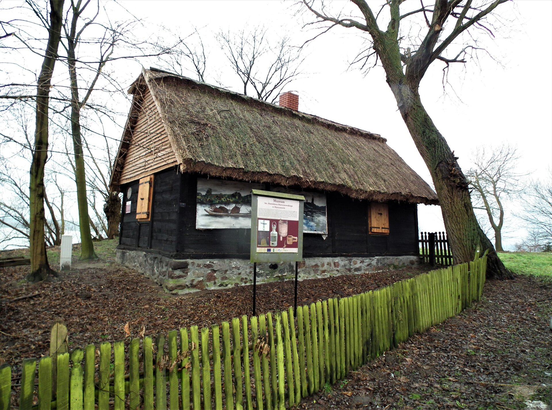 Fotografia przedstawia drewnianą chatę ze słomianym dachem ogrodzoną drewnianym płotem. Wokół chaty drzewa i tabliczka informacyjna.