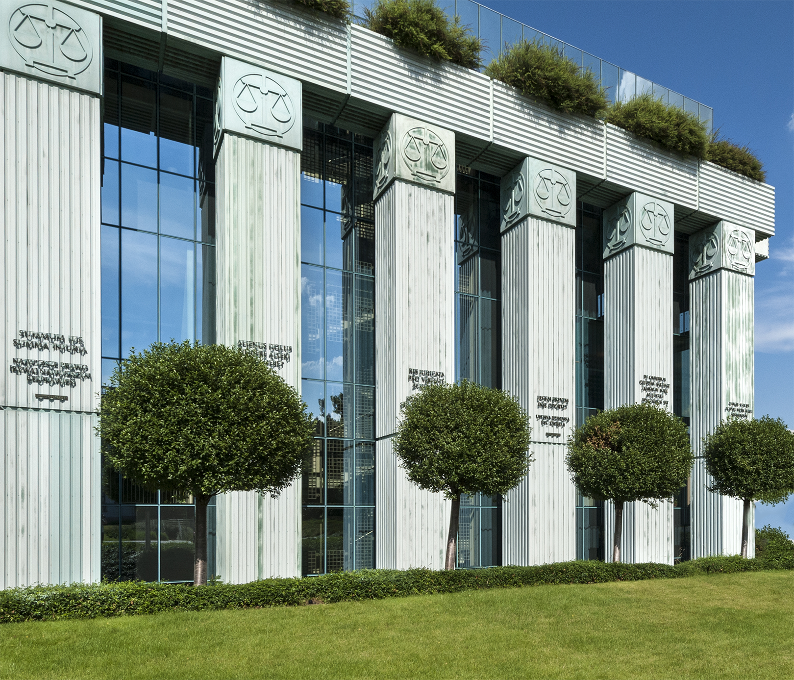 Zdjęcie przedstawia budynek z betonowymi kolumnami i za nimi wąskimi oknami. Na głowicach kolumn widnieje motyw wagi a na środku kolumn umieszczono paremie prawnicze ( krótkie sentencje). Nad szczytami  kolumn mur, na którym rośnie zieleń. Przed budynkiem rozciąga się żywopłot, trawa i drzewa uformowane w kule. 