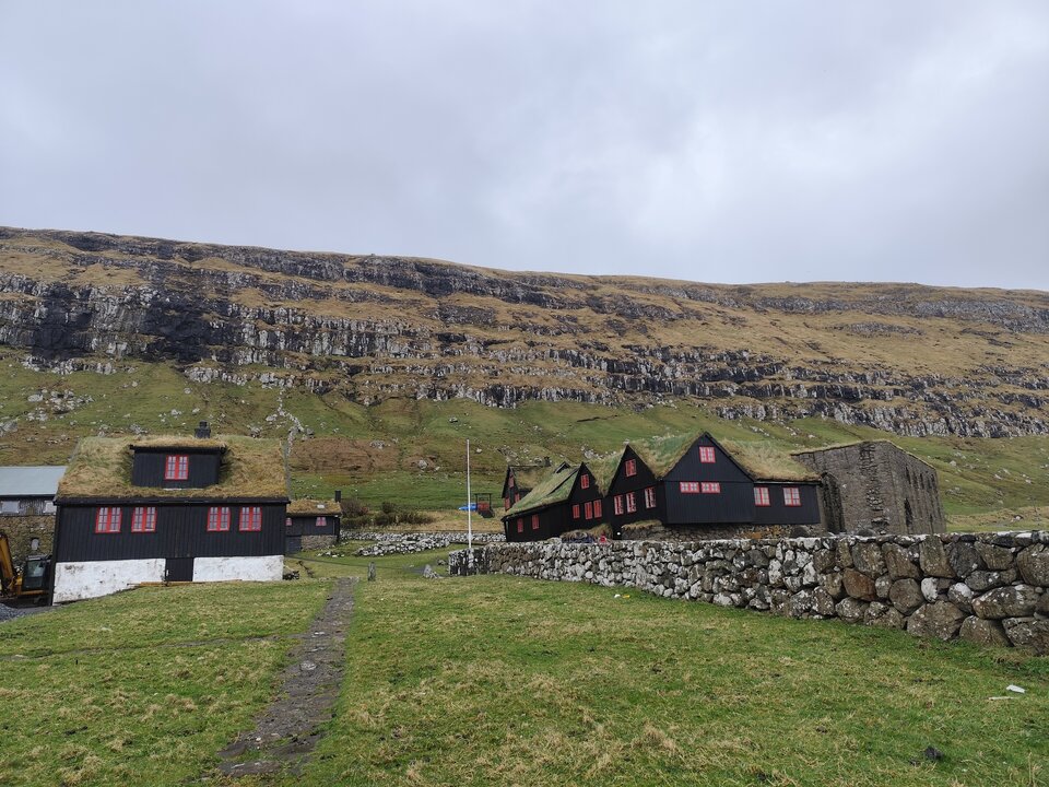 Kadr zdjęcia jest szeroki i pokazuje spokojny, surowy krajobraz. Na dole rozciąga się nierówna, zielona łąka; przez jej środek biegnie wąska ścieżka z ciemnych kamieni, która kieruje wzrok ku zabudowaniom. Po lewej stoi większy, prostokątny dom z czarnej deski z białym pasem przy ziemi; ma darniowy dach porośnięty trawą oraz kilka okien z czerwonymi ramami. Po prawej stronie widać kilka mniejszych domów ustawionych w szeregu, również czarnych, z zielonymi dachami z darni i czerwonymi oknami; część ma wysokie, trójkątne szczyty. Przed tą grupą domów ciągnie się kamienny mur z dużych, nieregularnych głazów. W tle znajduje się strome zbocze z wyraźnymi warstwami skał i plamami niskiej roślinności; na stokach widać rozrzucone kamienie. Niebo jest jednolicie zachmurzone, co wzmacnia wrażenie chłodu i wilgoci.