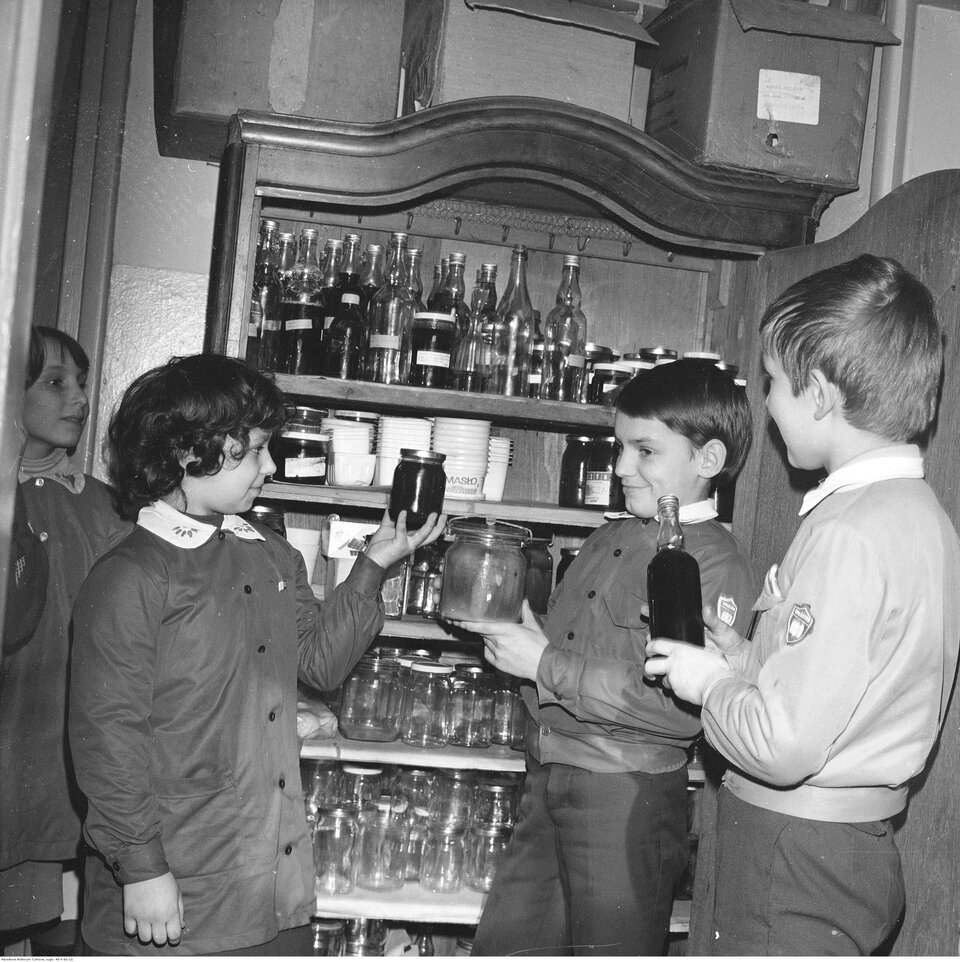 Fotografia przedstawiająca grupę dzieci, ubranych w mundurki szkolne. W dłoniach trzymają słoiki i butelki z przetworami.  Stoją przy otwartym kredensie. Na półkach kredensu stoją liczne butelki i słoiki z przetworami.