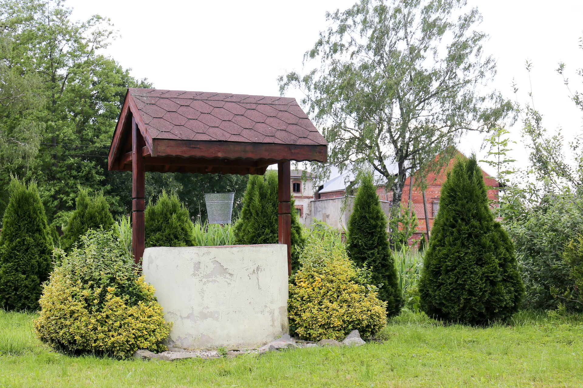 Fotografia przedstawia studnię umieszczoną w ogrodzie. Nad nią spadzisty daszek, dookoła krzewy i drzewa.