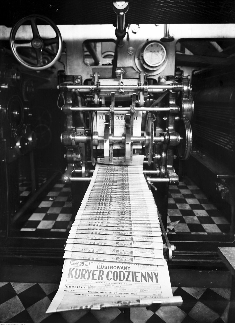 Centralnym punktem zdjęcia jest maszyna drukarska, z której wychodzą świeżo zadrukowane egzemplarze gazety. Na pierwszym planie widać układające się równo strony z tytułem: „Ilustrowany Kuryer Codzienny”. Maszyna pracuje pełną parą – rolki i elementy mechaniczne wskazują na przemysłową skalę produkcji. Tło stanowią metalowe i mechaniczne części prasy oraz czarno‑biała podłoga w szachownicę.