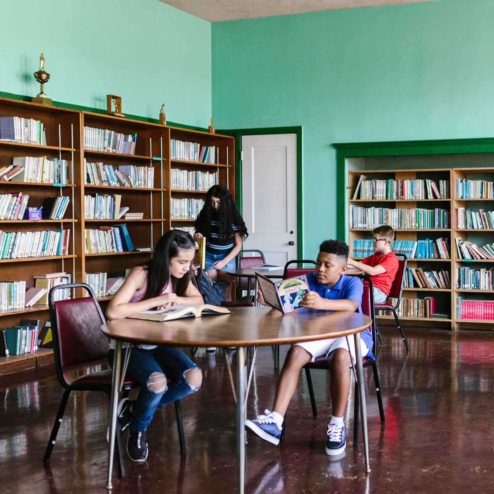 Na zdjęciu widzimy czwórkę dzieci w wieku szkolnym z książkami, przy stołach stojących we wnętrzu biblioteki. W tle duże regały wypełnione książkami.