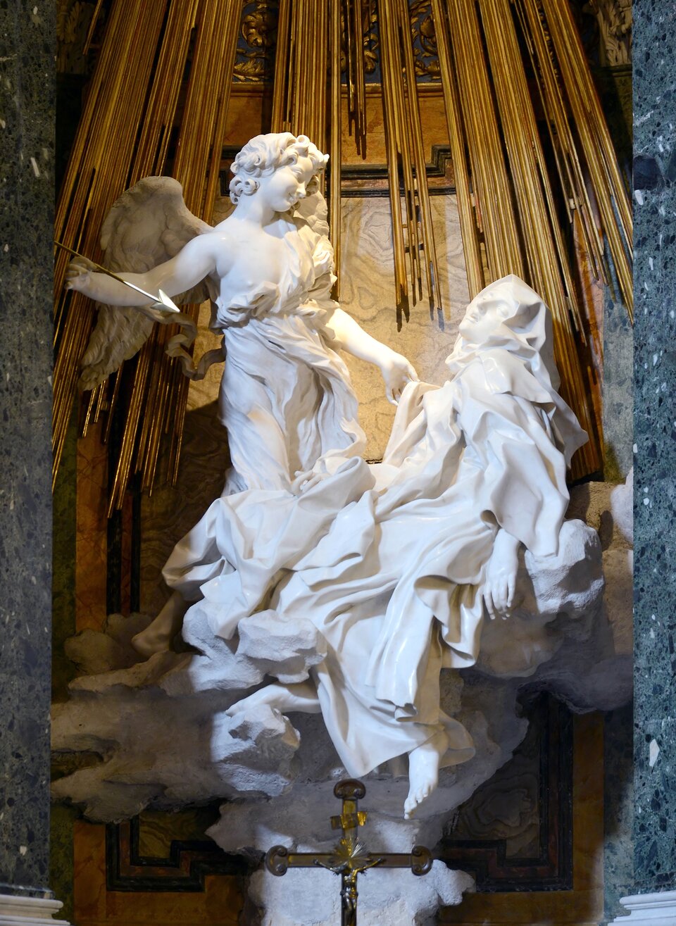 Na zdjęciu została ukazana rzeźba, z marmuru, świętej Teresy i anioła na obłoku. Kobieta ukazana jest w pozycji półleżącej, a anioł stojącej. Głowa świętej jest lekko przechylona w lewą stronę, do widza, ma ona półprzymknięte oczy i rozchylone usta, a jej lewa dłoń swobodnie opada wzdłuż ciała. Mocno udrapowana szata św. Teresy okrywa ją w całości, a jej całe ciało zwraca się w stronę strzały, którą trzyma w prawej dłoni stojący naprzeciw niej anioł i wymierza nią w postać świętej. Lewą dłonią dotyka jej szaty, przechylając lekko głowę w lewą stronę. Ma on kręcone, nieco dłuższe włosy, okryty jest lekko udrapowaną tkaniną, spod której widać prawy bark. Na dwie postacie z góry spływają liczne złote promienie. Poniżej rzeźby znajduje się krzyż.