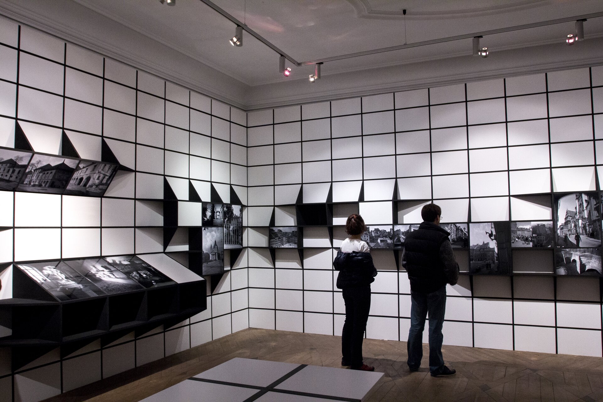 Na zdjęciu znajduje się pomieszczenie o białym suficie, brązowej podłodze z szaro‑czarnymi elementami oraz biało‑czarnych ścianach. Na ścianach są małe, prostokątne, białe płytki z czarnymi fugami. Na ścianach, na różnych wysokościach zamontowane są czarne wystające elementy, na których są biało‑czarne ilustracje. Na podłodze, po lewej stronie, są szare prostokąty z czarnymi liniami pomiędzy nimi. W pomieszczeniu znajdują się 2 postacie, które stoją tyłem. Na suficie wiszą szare szyny z małymi lampkami.  