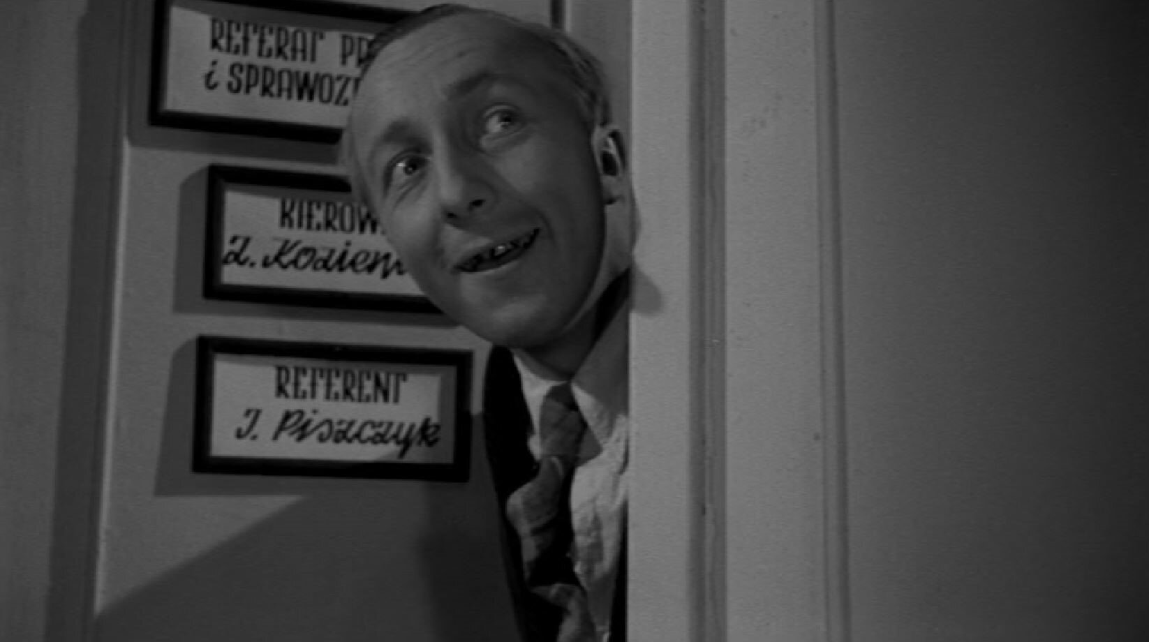Zdjęcie przedstawia głównego bohatera wyglądającego przez drzwi. Na drzwiach widnieją trzy tabliczki. jedna z nich jest z napisem referent J. Piszczyk.