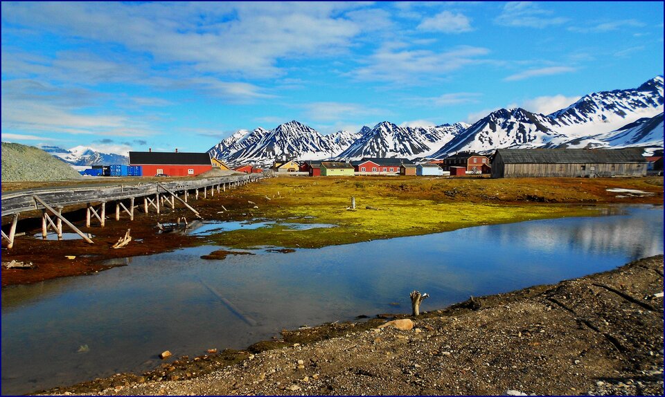 Zdjęcie przedstawia osadę Ny‑Ålesund. Na pierwszym planie widoczny jest akwen wodny, który przecina niewielki most na drewnianych podporach. W osadzie dominują niskie budynki mające czerwone ściany oraz spadziste dachy. Za osadą są wzgórza pokryte śniegiem.