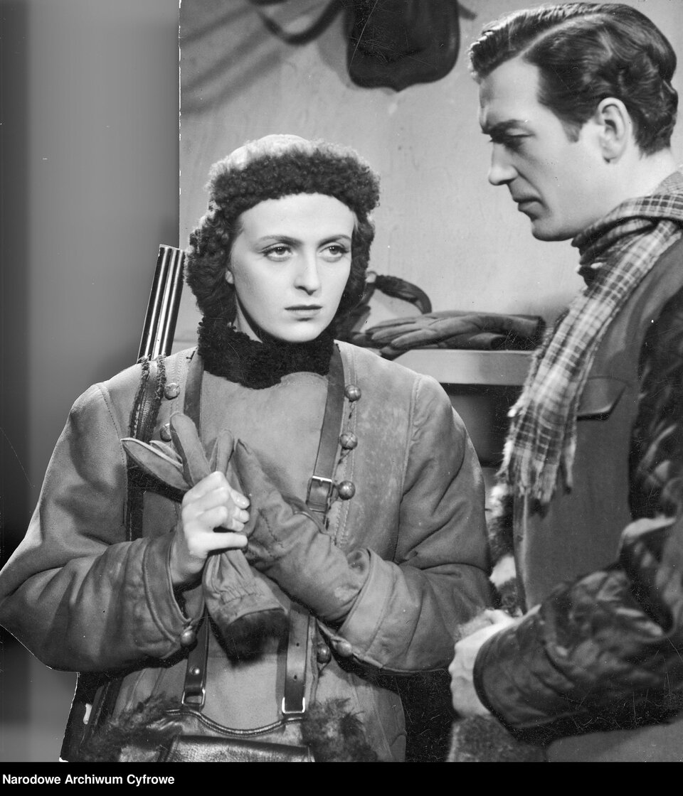 Fotografia przedstawiająca dwoje młodych ludzi. Po lewej stronie stoi kobieta. Ukazuje prawy półprofil. Zaprezentowana jest od pasa w górę. Kobieta ma owalną twarz, wyraźnie zarysowane brwi, duże oczy i prosty nos. Na głowie ma czapkę. Spod niej wystają półdługie, kręcone włosy. Ubrana jest w kurtkę. Z dłoni zdejmuje skórzane rękawiczki. Przez szyję ma przewieszoną skórzaną torbę. Na lewym ramieniu wisi pasek, do którego przymocowano strzelbę. Po prawej stronie stoi mężczyzna. Postać ukazuje lewy profil. Ma owalną twarz. Jego włosy są krótkie, zaczesane na prawą stronę, Delikatnie marszczy wyraziste brwi. Jego nos jest niewielki i prosty. Patrzy na kobietę. Mężczyzna jest ubrany w pikowaną kurtkę i kamizelkę. Szyja mężczyzny jest okręcona szalikiem w kraciasty wzór. W tle znajduje się ściana, na której wisi półka. Na półce leży para męskich, skórzanych rękawiczek.