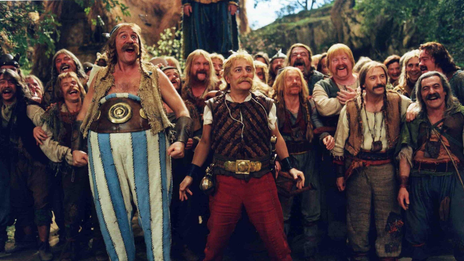 Na zdjęciu znajduje się grupa mężczyzn stojących oraz śmiejących się, mężczyźni patrzą naprzeciw siebie. Na pierwszym planie znajdują się Asterix i Obelix. Asterix ubrany jest w brązowa koszulkę oraz czerwone spodnie. Obelix ubrany jest w niebiesko białe spodnie w paski z ogromnym paskiem oraz brązową kamizelkę. Na drugim planie znajduje się tłum mężczyzn. Mężczyźni ubrani są w spodnie, koszule i kamizelki. W tle ilustracji znajduje się las oraz wzniesienia. 