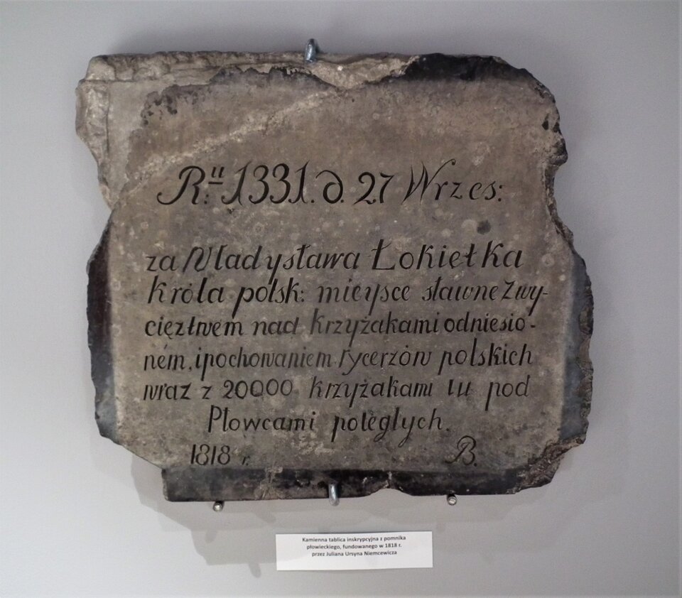Na fotografii widnieje kamienna płyta o nierównych krawędziach. Na płycie widnieją napisy w języku polskim. Poniżej płyty znajduje się opis muzealny umieszczony na białej kartce, przyklejonej do ściany poniżej eksponatu. 