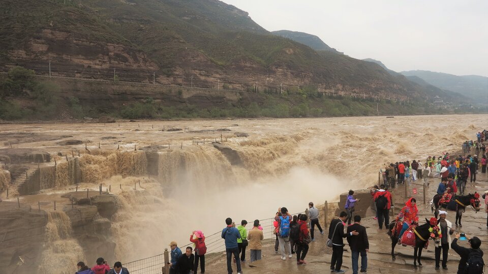 Na zdjęciu jest szeroka rzeka, której woda jest wzburzona. Ilość wody jest imponująca. Woda ma kolor żółty, po lewej stronie zdjęcia spływa po skalnych stopniach. Na pierwszym planie na tarasie rzekę obserwują turyści, robią zdjęcia. Lewy brzeg rzeki stanowią wzniesienia. 