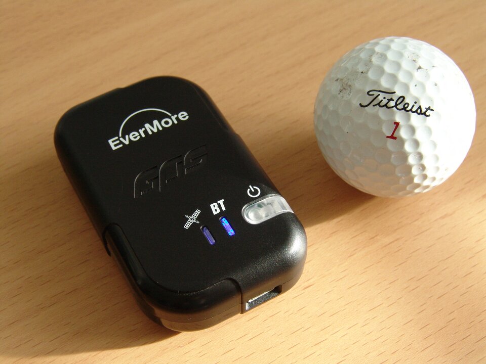 Na zdjęciu znajduje się rejestrator danych GPS. Jest to małe urządzenie w formie czarnego, plastikowego, spłaszczonego prostopadłościanu. Widoczne są trzy diody - jedna oznaczona ikonką satelity, druga literami B T, trzecia symbolem włącznika. Na opakowaniu widnieje napis Evermore. Obok urządzenia znajduje się piłeczka golfowa, jest o połowę mniejsza od rejestratora. 