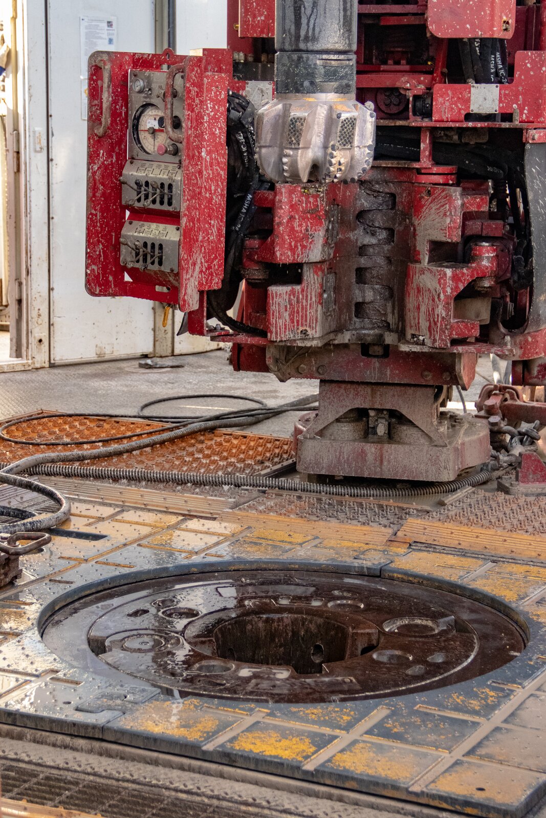 Na zdjęciu przedstawiono stół wiertniczy. W dolnej części zdjęcia widoczny jest fragment posadzki. Na posadzce znajduje się żółty metalowy prostokąt. W prostokąt wpisany jest okrąg. W środku okręgu widoczny jest otwór. W tle widać rurę wiertnicza na końcu której znajduje się koronka.  