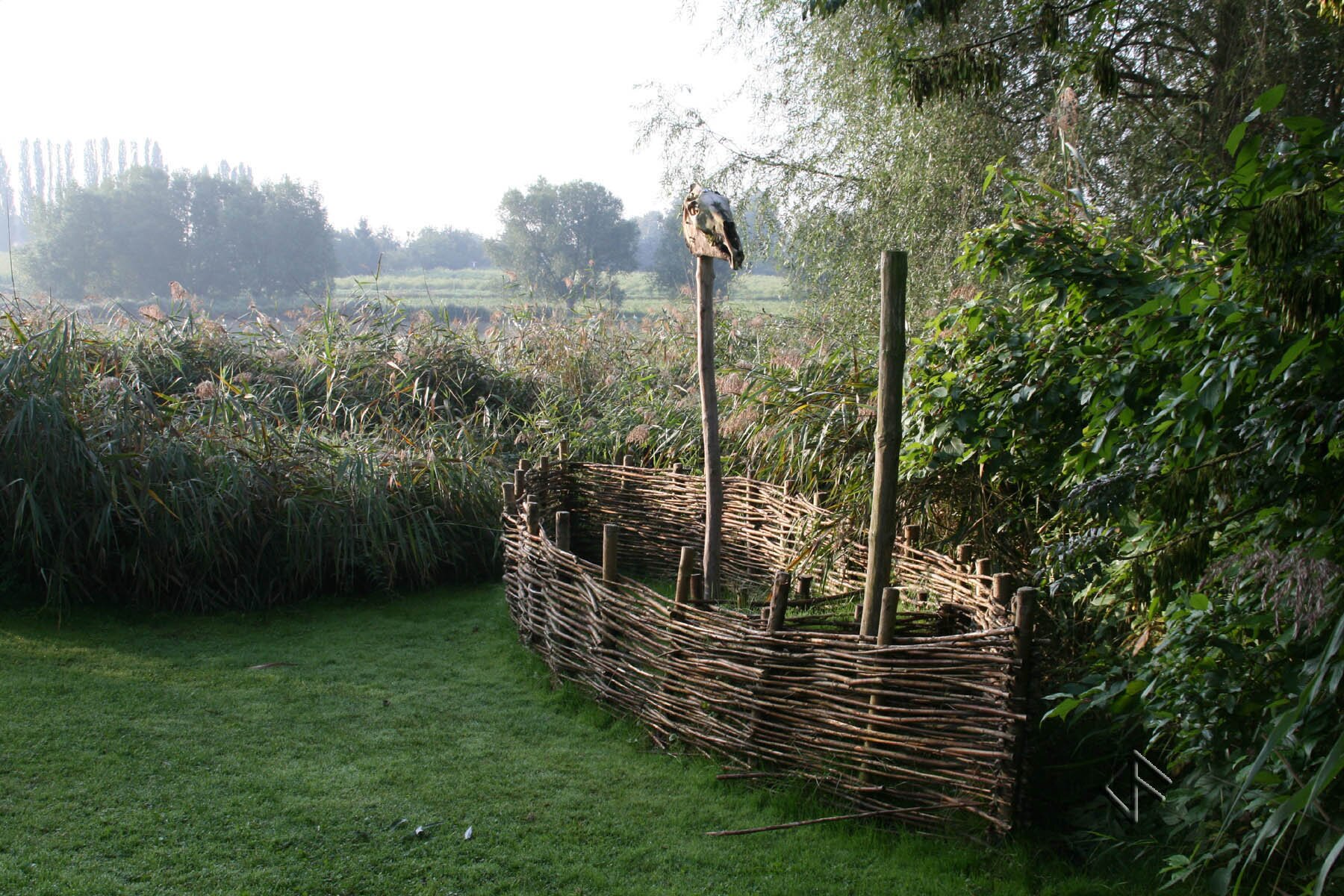 Zdjęcie współczesne, przedstawiające rekonstrukcję miejsca kultu. Złożona z drewnianych gałązek konstrukcja w kształcie łodzi, oparta o drewniane paliki z wewnętrznej strony, stoi na trawie bliżej prawej krawędzi zdjęcia. Z wnętrza łodzi wystają dwie drewniane, wysokie belki. Na jednej z nich umieszczono czaszkę dużego zwierzęcia hodowlanego - prawdopodobnie konia lub krowy. W głębi ilustracji widoczne sitowia i zadrzewione pole, przy lewej krawędzi ilustracji trawnik, po prawej stronie zdjęcia gęste zadrzewienie skraju zagajnika. 