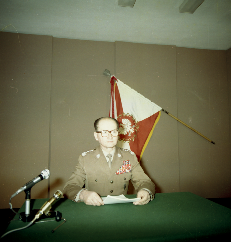 Kliknij, aby powiększyć Na zdjęciu przedstawiono starszego mężczyznę. Jest łysiejący, nosi okulary. Ubrany jest w mundur z odznaczeniami. Siedzi przy stole. W rękach trzyma kartki. Za nim flaga Polski z orłem z okresu PRL, przed nim mikrofony stojące na stole.