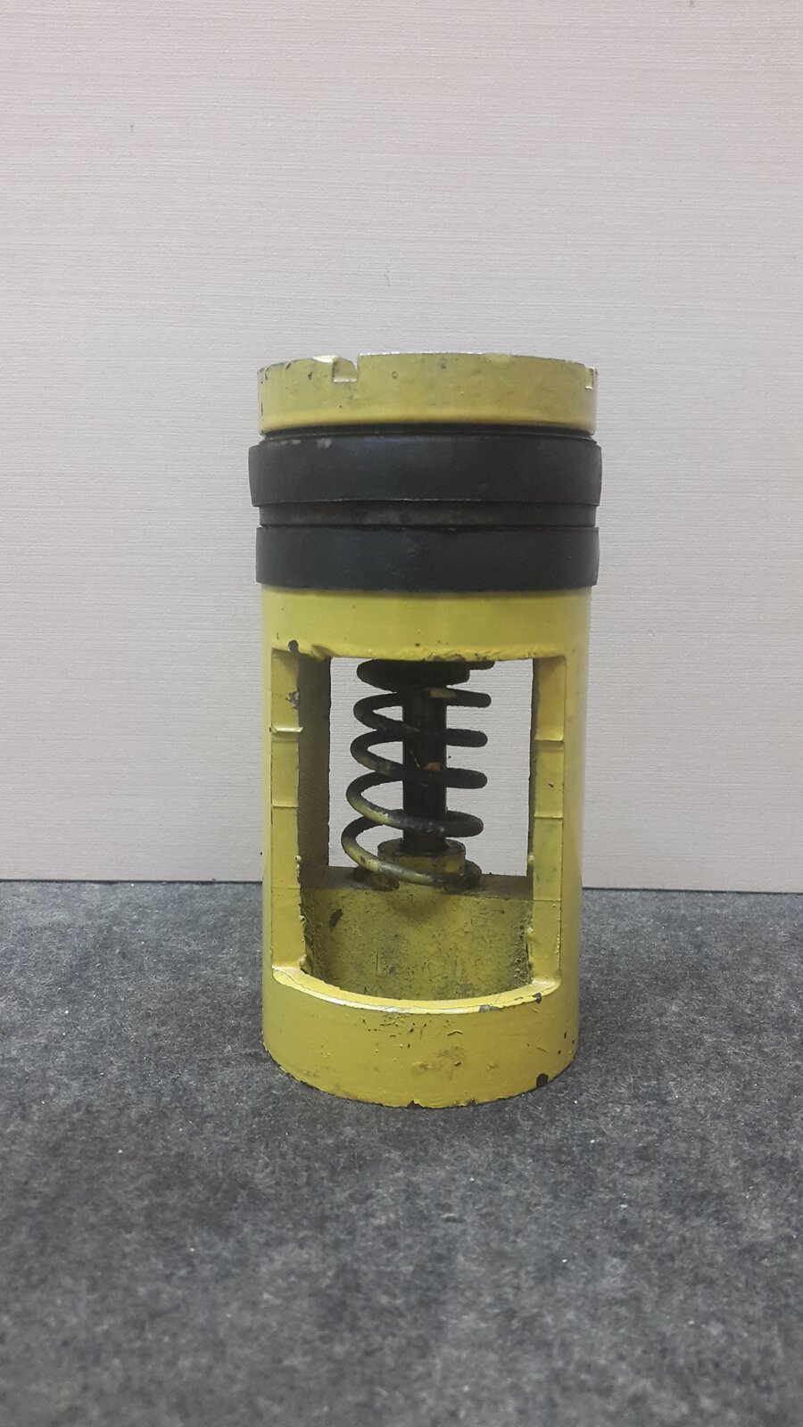  Na zdjęciu widoczny jest wrzutowy zawór zwrotny. Przedstawiony  zawór jest w formie walca. Na górną cześć walca zamontowany jest czarny gumowy pierścień. Dolna część walca składa się z dwóch ścian bocznych. Pozostała przestrzeń nie ma wypełnienia. Przez otwór w ścianie widać stalowy pionowy rdzeń. Wokół rdzenia nawinięta jest gruba sprężyna. Rdzeń łączy się z żółtą podstawą walca.Poniżej zdjęcia w prawym dolnym rogu widoczny jest przycisk odtwarzania. Tekst nagrania jest zgodny z tytułem zdjęcia.