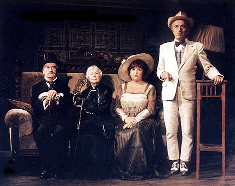 Na zdjęciu widać postacie pozujące do zdjęcia. Trzy z nich siedzą na jasnobrązowej starodawnej kanapie.  Z lewej strony siedzi starszy mężczyzna w czarnym garniturze i cylindrze. Opiera się o laskę, trzymając w dłoniach białe rękawiczki. Obok niego siedzi starsza kobieta w czarnej starodawnej czarnej sukni, która  sięga jej do kostek. Włosy ma upięte, ręce opiera na czarnej lasce. Obok niej po prawej stronie kanapy siedzi kobieta w wieku 50 lat. Ma na głowie przekrzywiony kremowy kapelusz z dużym rondem, ubrana jest w kremową suknię z dużym prostokątnym dekoltem. Obok niej stoi elegancko ubrany mężczyzna. Ma na sobie kremowy garnitur, brązową muszkę, kamizelki w kratkę i kapelusz. Lewą rękę opiera o komodę.