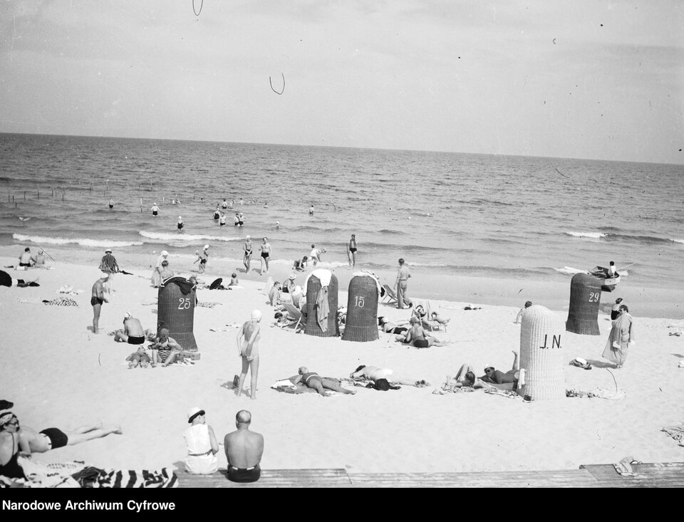 Zdjęcie przedstawia plażę nad morzem. Ludzie odpoczywają i kąpią się w morzu. Na plaży przebywa wielu ludzi (plażowiczów), zarówno w wodzie jak i wypoczywających na piasku. Wśród nich są kobiety, mężczyźni i dzieci w strojach kąpielowych z lat 30. Widać kosze plażowe wykonane z wikliny.