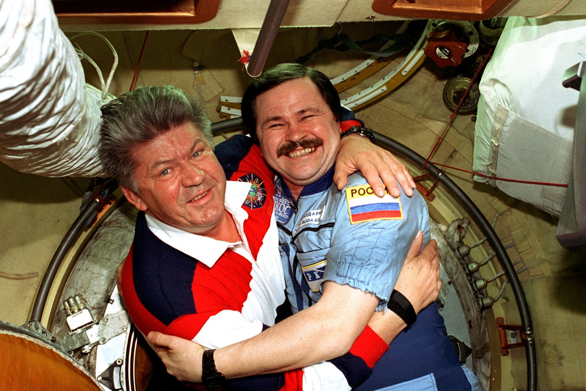 Zdjęcie przedstawia dwóch kosmonautów na stacji Mir. Mężczyźni w wieku ok. 50 lat. Obejmują się i pozują do zdjęcia. Mężczyzna po lewej ma siwiejące włosy, ogorzałą twarz oraz widoczne zmarszczki. Oczy jasne. Nos duży. Ubrany w koszulkę polo z długim rękawem w granatowo‑czerwono‑białe pasy z białym kołnierzykiem. Mężczyzna po prawej ma krótkie, ciemne włosy, starannie zaczesane z przedziałkiem po prawej stronie (jego lewej). Mężczyzna szeroko się uśmiecha pokazując zęby. Oczy zmrużone. Mężczyzna ma ciemne wąsy. Ubrany jest w niebieski kombinezon z naszytą na lewym ramieniu flagą Rosji. Obaj mężczyźni noszą czarne zegarki.