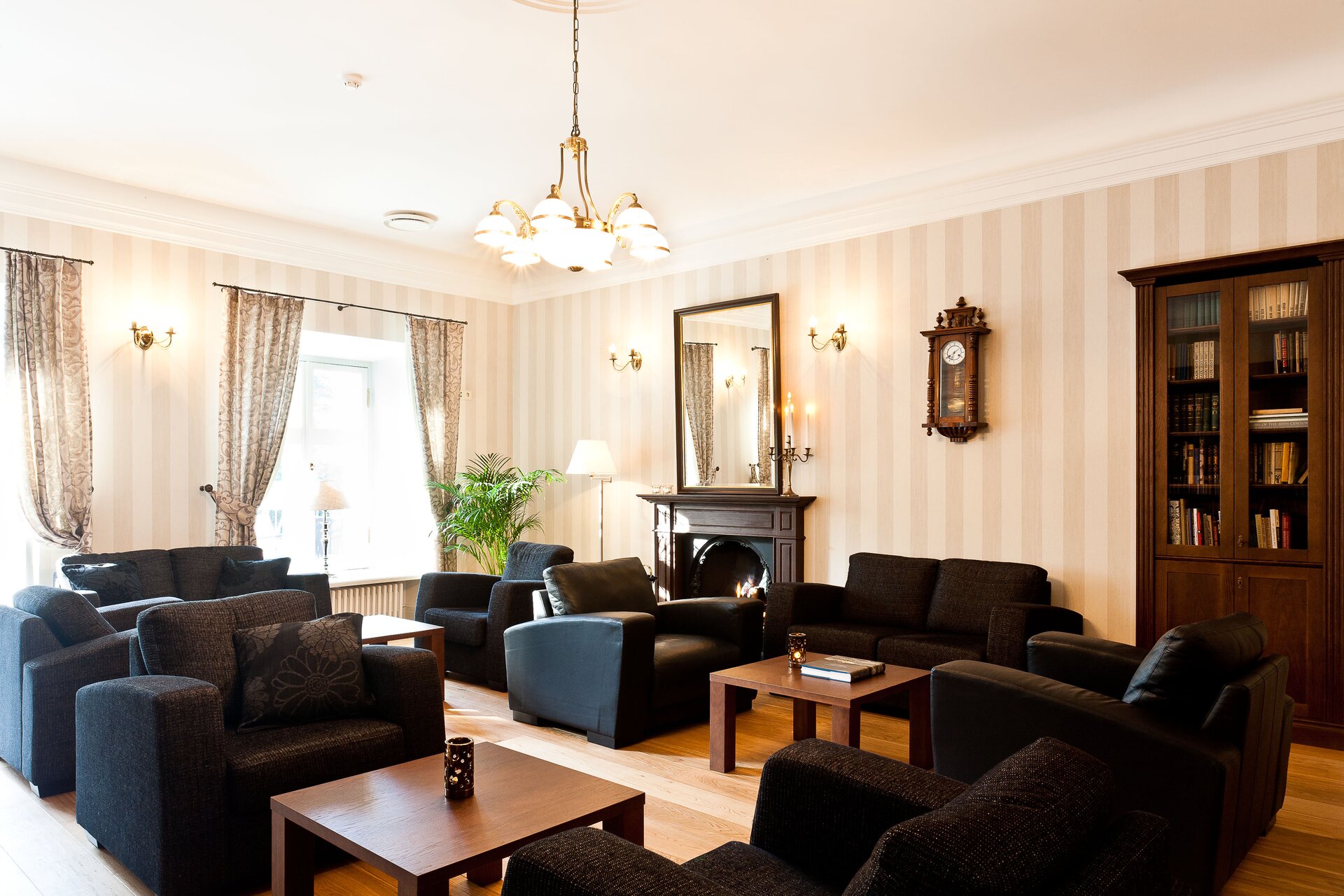 Zdjęcie przedstawia przestronne pomieszczenie. Jego ściany wyłożone są tapetą w kremowe pionowe pasy. Na środku pomieszczenia znajdują się liczne czarne fotele z poduchami i sofy oraz proste stoliki pomiędzy nimi. Na stoliku stojącym bliżej stoi owalna ozdoba, zaś na stoliku stojącym nieco dalej leży książka. Pod ścianą po prawej stronie zdjęcia stoi przeszklony regał z książkami, a obok wisi stylowy drewniany zegar antyczny z rzeźbieniami i białą tarczą. Na lewo od zegara znajduje się kominek z wiszącym nad nim lustrem. Na ścianie po lewej stronie zdjęcia znajdują się dwa duże okna z zasłonami w delikatny wzór. W pomieszczeniu są zapalone lampy: sufitowa i stojąca przy kominku. Podłoga jest brązowa i drewniana.