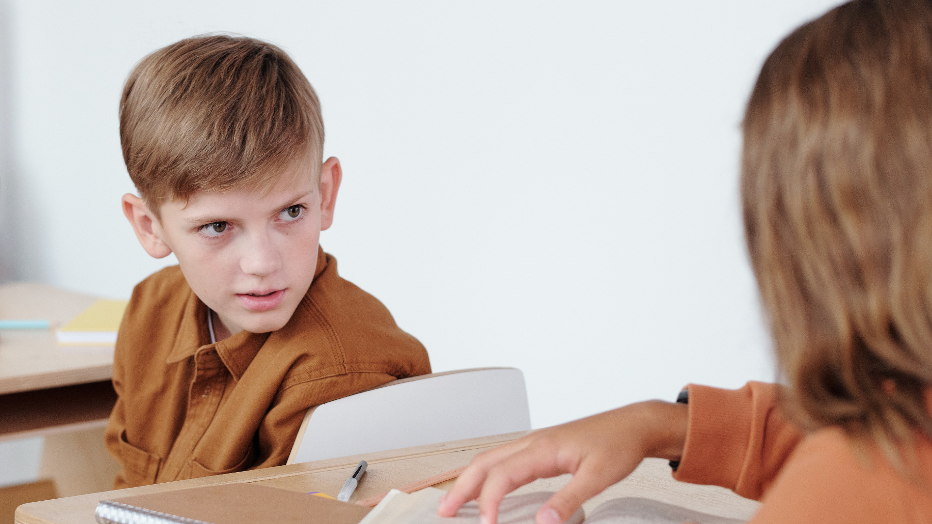 Zdjęcie przedstawia chłopca i dziewczynkę siedzących w ławkach szkolnych. Chłopiec odwraca się do dziewczynki, która siedzi w ławce za nim. Niewidoczna jest twarz dziewczynki.