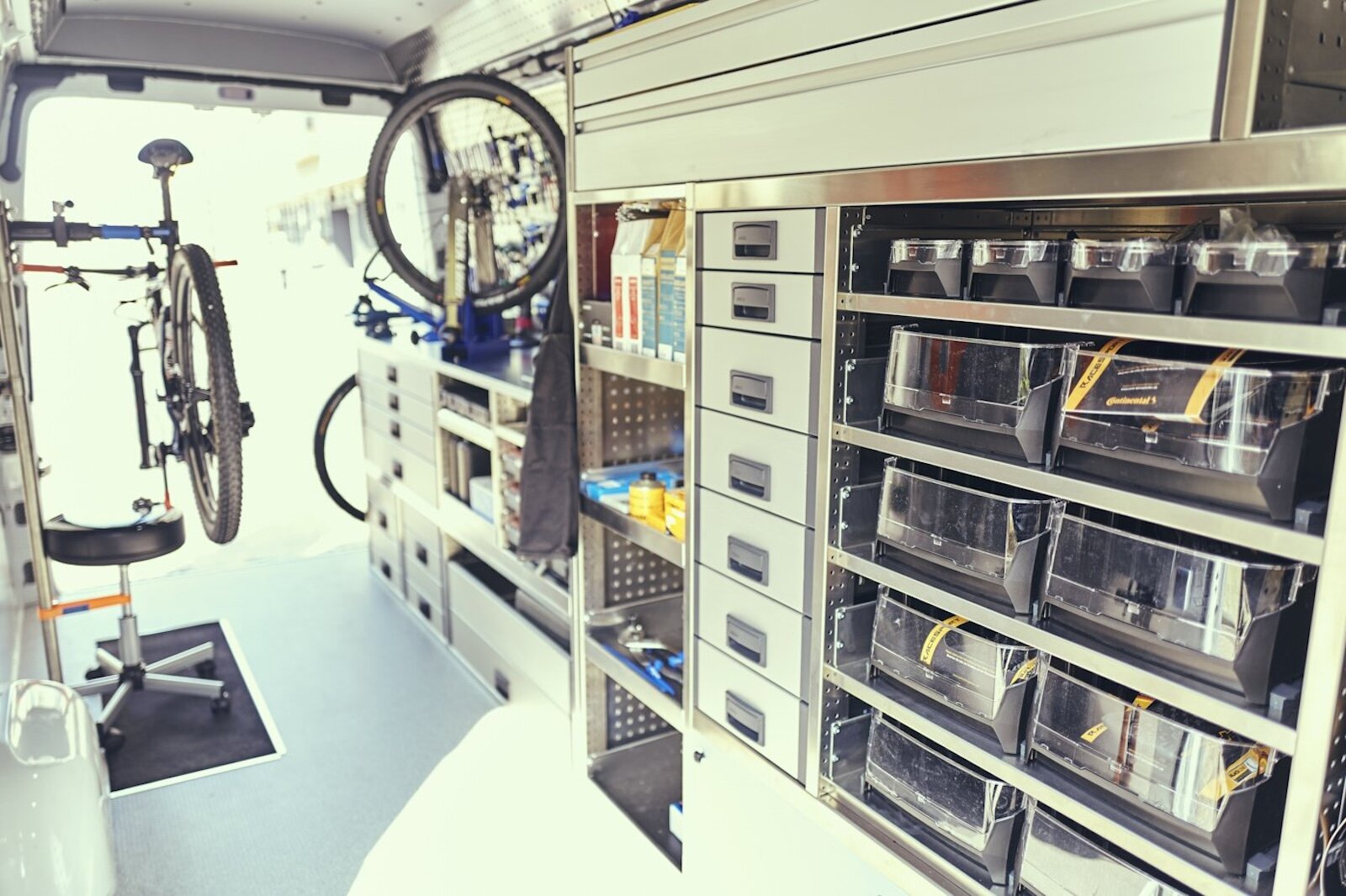 Zdjęcie przedstawia wnętrze furgonetki, w której znajdują się pudełka z narzędziami i częściami zamiennymi do rowerów. 