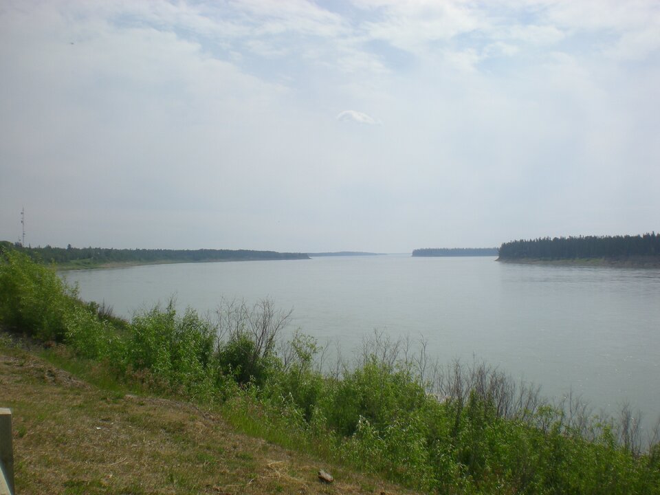  Zdjęcie wykonane z brzegu rzeki. W pierwszym planie niska roślinność, dalej rozległa tafla spokojnej wody. Tło rozmywa się w delikatnej mgle. Kolory stonowane, dominują szarości, zielenie i błękity. Rzeka szeroka, powolna, płynąca przez równinny krajobraz. Po obu stronach niska roślinność. Brak widocznych zabudowań. 