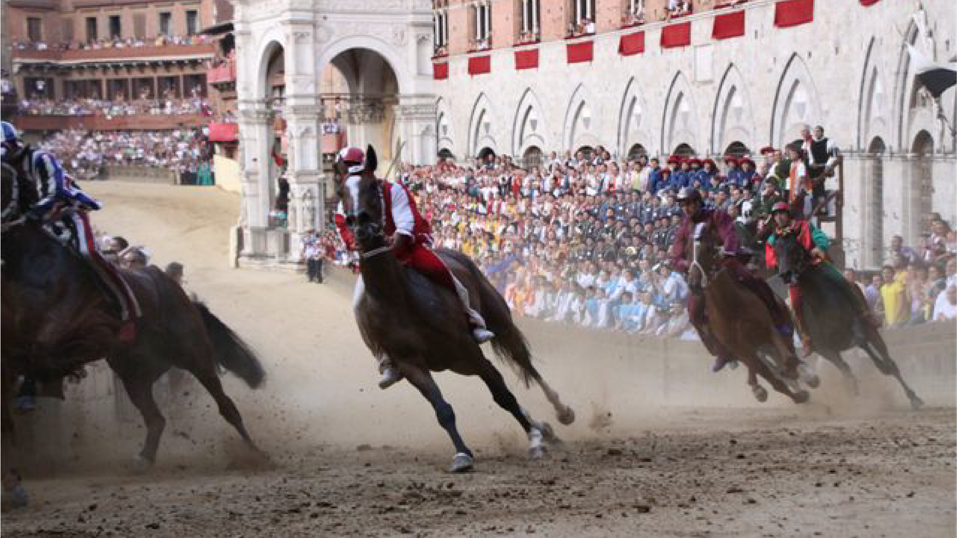 Zdjęcie przedstawia gonitwę konną na głównym placu Sieny Piazza del Campo trakcie festynu Palio. Jężdźcy ubrani są w kolorowe kostiumy z epoki, które reprezentują dzielnice Sieny. Na pierwszym planie biegną cztery konie. Po prawej stronie na trybunach ustawionych na tle starych murów siedzą widzowie.